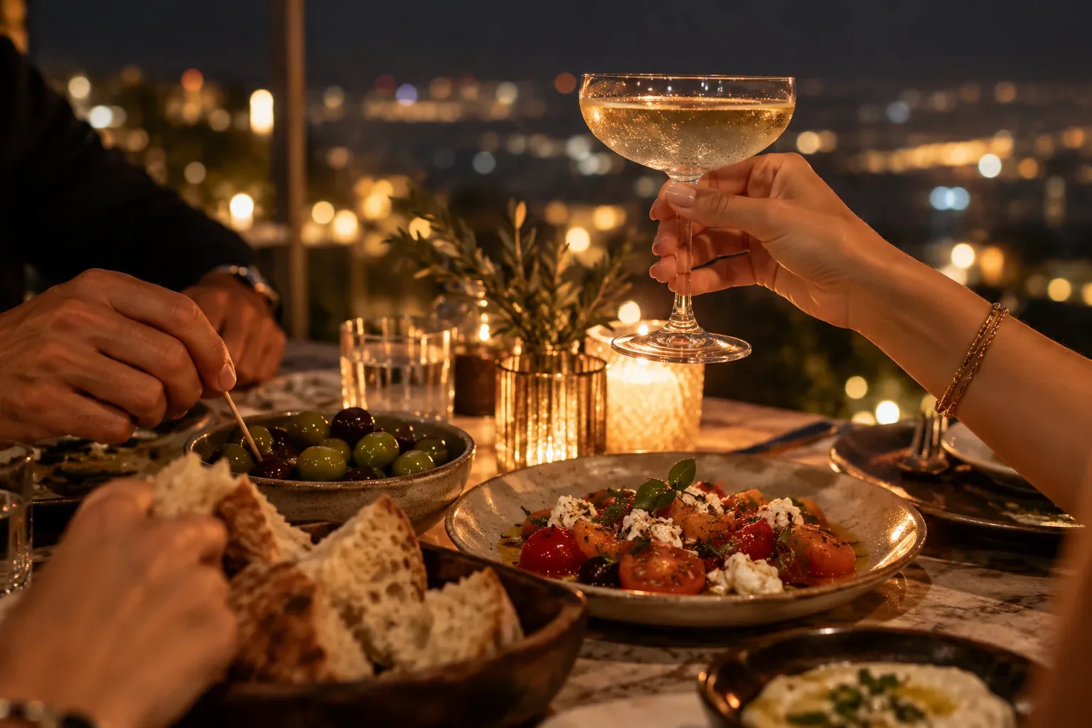 A table of friends with a coupe, olives and tomatoes above the city at night